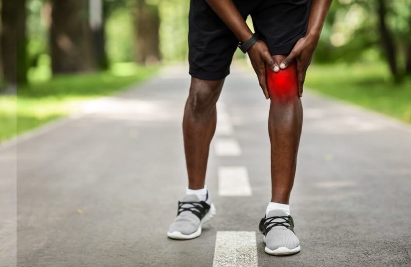 Cropped of black sportsman touching his inflamed knee highlighted with red, park background, copy space